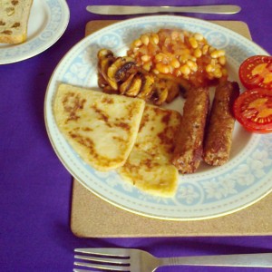 Plate with 2 potato scones, 2 vegan sausages, a clump of beans in tomato sauce, 2 tomato halves, fried sliced mushrooms, on a purple tablecloth with the corner of some toasted fruit bread showing